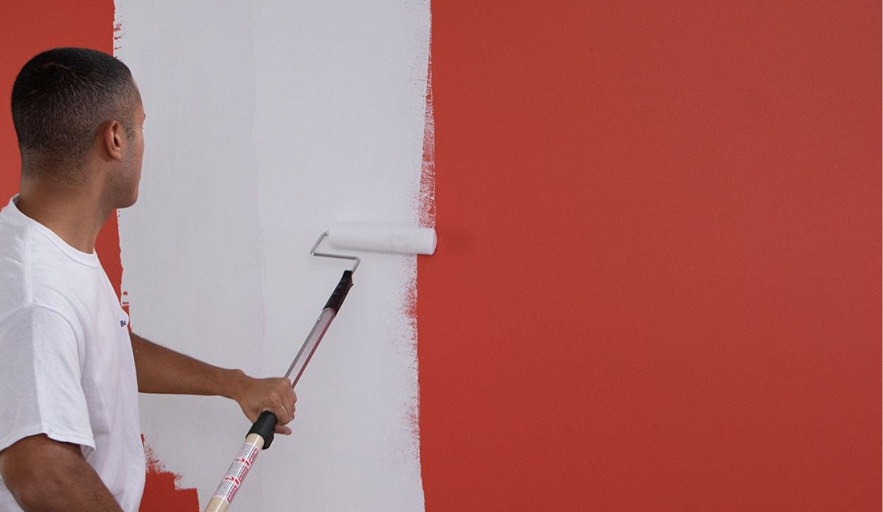 Painter using a roller brush to apply white primer on a bright red wall